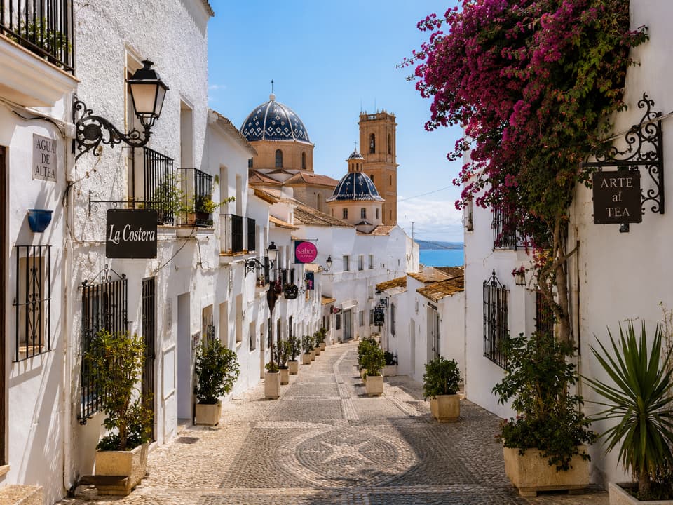 Calle empedrada del casco antiguo de Altea con casas encaladas, buganvilla y la cúpula azul de la iglesia al fondo
